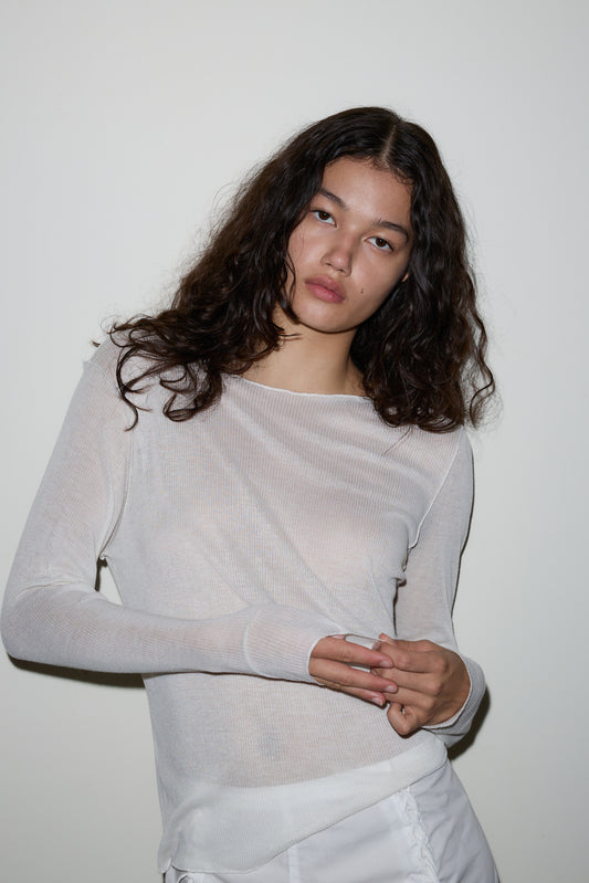 A woman with long, wavy dark hair wears a sheer white Knit Long Sleeve and white pants, standing against a plain white wall and looking directly at the camera with a neutral expression.