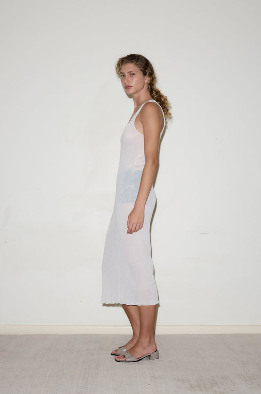 A person with curly hair stands sideways against a plain white wall, wearing the Knit Singlet Dress - Off White and open-toed heeled sandals.