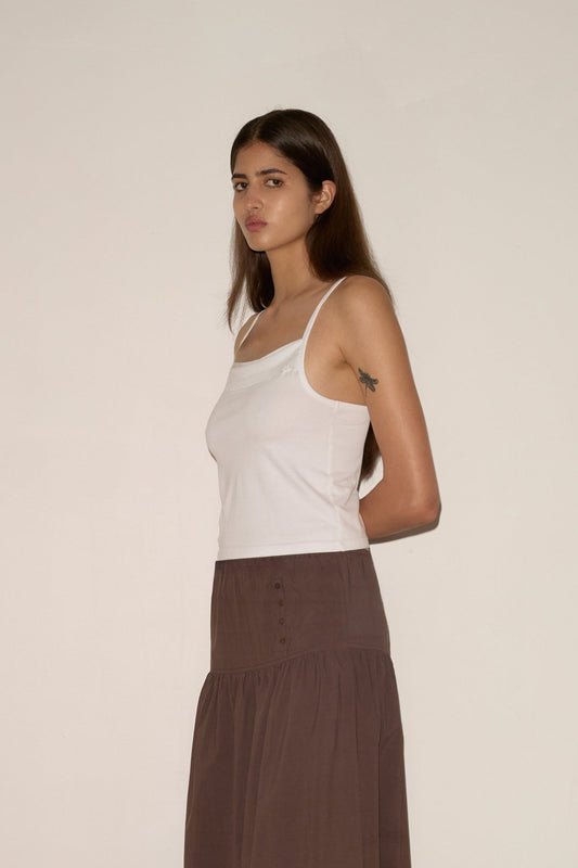 A woman with long brown hair stands sideways against a white wall, wearing a white sleeveless top, the 0 Button Skirt - Chocolate, and beige flats. Her hands are behind her back.