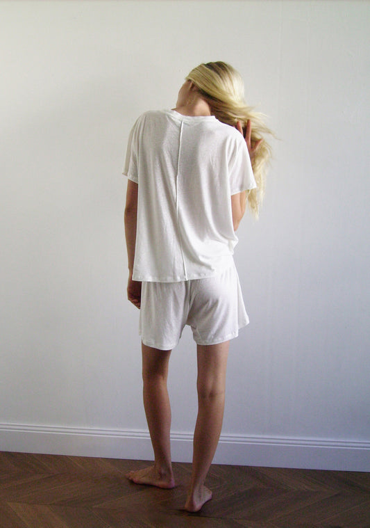 A person with long blonde hair, wearing 0s soft short - ecru and a loose white tee, stands barefoot on a wooden floor facing away from the camera, gently touching their hair against a plain white wall.