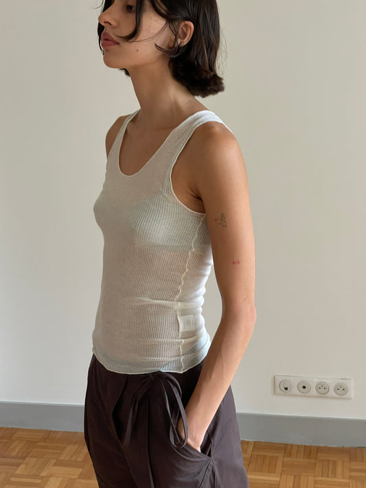 A person with short dark hair wears the Knit Tank - Off White, made from eco-friendly fine knit fabric, paired with dark pants. Indoors against a plain wall with outlets, their left arm displays a small tattoo and faint red mark.