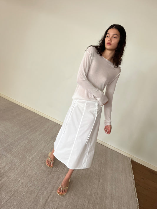 A woman with long dark hair, barefoot on a light rug against a plain wall, wears the Knit Long Sleeve - Off White with a white skirt, looking slightly to the side.