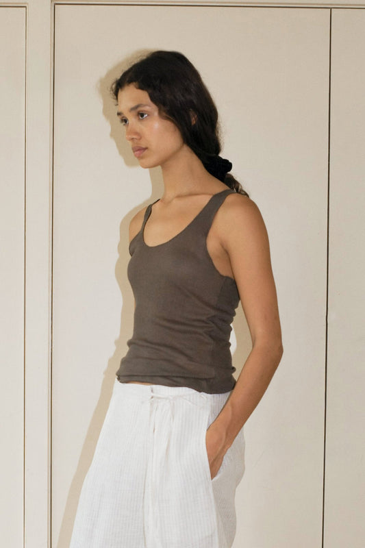 A person with long dark hair wears a Knit Tank and loose white pants, standing indoors with one hand in their pocket and gazing thoughtfully to the side.